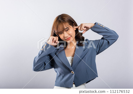 An Asian woman is captured in a studio shot, conveying her stress and discomfort caused by loud noise. She covers her ears and closes her eyes tightly, isolated on a white background. An Asian woman is captured in a studio shot, conveying her stress and discomfort caused by loud noise. She covers her ears and closes her eyes tightly, isolated on a white background. 112176532