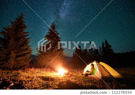 Night camping in mountains under starry sky and Milky way. Female tourist sitting in tent entrance in campsite, admiring landscape and burning campfire. Concept of traveling and hiking. Night camping in mountains under starry sky and Milky way. Female tourist sitting in tent entrance in campsite, admiring landscape and burning campfire. Concept of traveling and hiking. 112176554