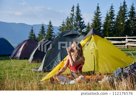 Woman hiker camping outdoors. Tourist sitting in campsite, admiring landscape and drinking tea. Sporty, young female having break, enjoying in mountains. Concept of beauty of nature. 112176576
