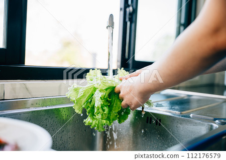 Hygienic food prep, Woman's hands washing fresh vegetables under running water in a modern kitchen sink for a vegan salad. Clean and fresh leafy greens for homemade healthy eating. 112176579