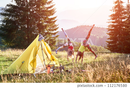 Two women hikers camping outdoors. Sporty females training, doing salto in campsite. Slim, young women traveling, hiking in mountains, enjoying nature. Concept of healthy lifestyle. 112176580