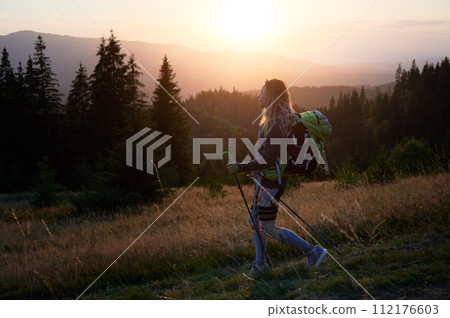 Woman hiker hiking outdoors at sunset. Sporty, positive woman traveling in mountains. Young, sporty female with backpack using trekking sticks, walking. Concept of harmony with nature. 112176603