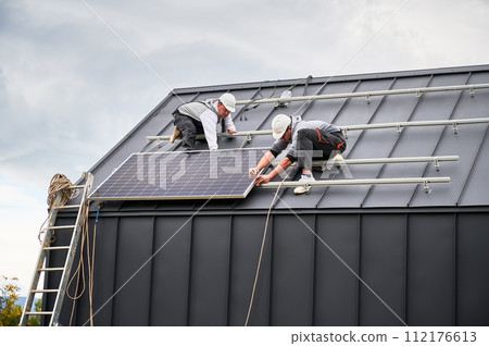 Mounters installing photovoltaic solar panels on roof of house. Men engineers in helmets building solar module system with help of hex key. Concept of alternative, renewable energy. 112176613