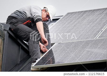 Man technician mounting photovoltaic solar panels on roof of house. Engineer in helmet installing solar module system with help of hex key. Concept of alternative, renewable energy. 112176615