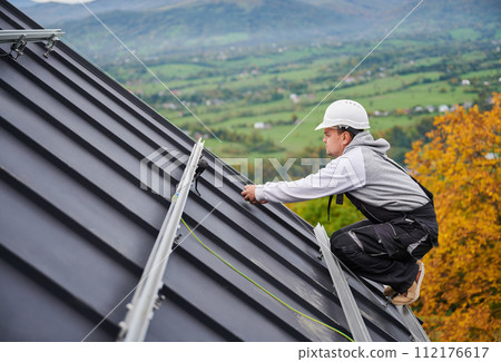Man roofer prepearing for mounting photovoltaic solar modules on roof of house. Electrician in helmet installing solar panel system outdoors. Concept of alternative and renewable energy. 112176617