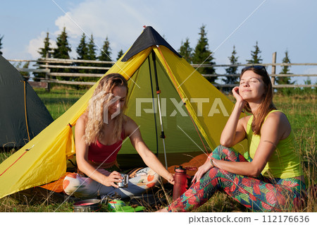Two women tourists camping in mountains. Attractive, positive women hiking in summer. Female friends sitting on grass near tent, having break. Concept of tourism and adventure. 112176636