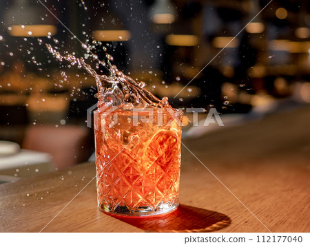Orange or red drink with splash and droplets and orange slice garnish on bar counter. Red brown cocktail with splashes in pub or nightclub or restaurant 112177040