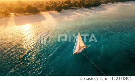 A sailboat over aquamarine waters, beneath golden light, near a golden sandy beach with palms in the background. 112178494