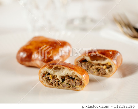 Small russian pies with minced meat Russian Piroshki. Cooked meat hand portioned pies or empanadas cut in half on white plate in elegant restaurant interior. Selective focus, close up. 112182186