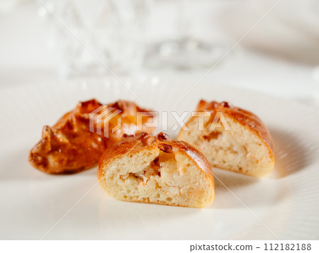 Small russian pies with minced salmon fish Russian Piroshki. Cooked tuna fish hand portioned pies or empanadas cut in half on white plate in elegant restaurant interior. Selective focus, close up. 112182188