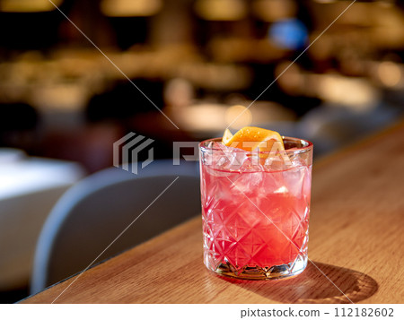 Transparent pink alcohol cocktail with ice, aperol and gin on bar counter in restaurant interior. Alcohol cocktail decorated orange zest spiral. Cold alcoholic cocktail in bar or nightclub. Copy space Transparent pink alcohol cocktail with ice, aperol and gin on bar counter in restaurant interior. Alcohol cocktail decorated orange zest spiral. Cold alcoholic cocktail in bar or nightclub. Copy space 112182602
