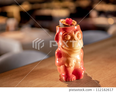 Authentic Tiki Drink Cocktail. Beautiful tropical tiki cocktail on bar counter. Tiki cocktail in monkey mug decorated fig and raspberry on bar in night club or restaurant interior 112182617
