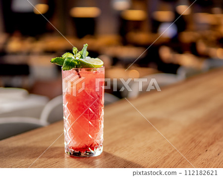 Red non-alcohol cocktail with crushed ice in glass on wooden bar counter in restaurant interior. Gin alcohol cocktail decorated lime and mint. Pink cold alcoholic cocktail in bar or nightclub 112182621