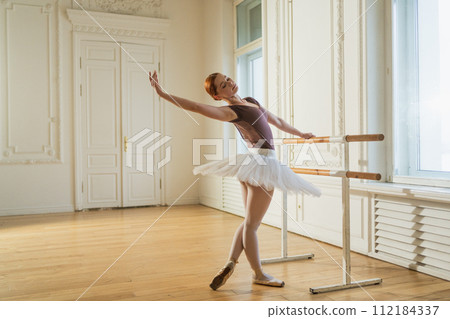 Young ballerina in tutu skirt stretching practicing ballet positions near ballet barre in dance class. Young classical dancer woman in dance studio. Ballet class training. Ballet school concept 112184337