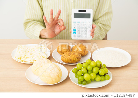 A middle-aged woman holding a food containing a lot of sugar (carbohydrates) and a calculator and making a circle sign A middle-aged woman holding a food containing a lot of sugar (carbohydrates) and a calculator and making a circle sign 112184497