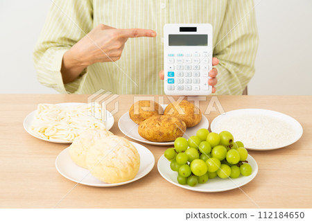 中年婦女指著含有大量糖(碳水化合物)的食物和計算器 中年婦女指著含有大量糖(碳水化合物)的食物和計算器 112184650