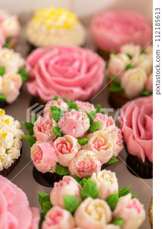 Encased in a pristine white paper cupcake box, each gourmet cupcake is a work of art, adorned with buttercream frosting flowers beautifully designed to resemble vibrant roses and tulips. 112185613