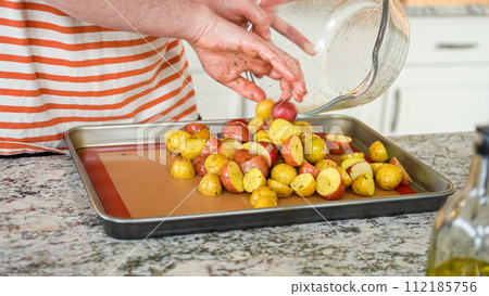 In a modern, white kitchen, a young man is engrossed in dinner preparations. His current endeavor includes arranging seasoned rainbow potatoes on a baking sheet, a meticulous step towards a 112185756