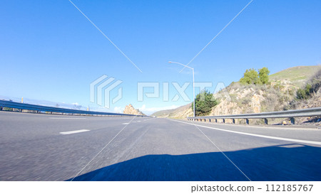 On a crisp winter day, a car cruises along the iconic Highway 1 near San Luis Obispo, California. The surrounding landscape is brownish and subdued, with rolling hills and patches of coastal 112185767