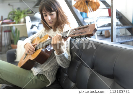 a woman plays her guitar while seated comfortably on a leather couch 112185781