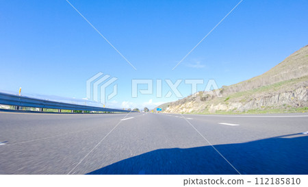 On a crisp winter day, a car cruises along the iconic Highway 1 near San Luis Obispo, California. The surrounding landscape is brownish and subdued, with rolling hills and patches of coastal 112185810