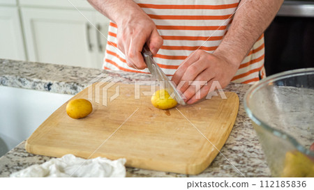 In the contemporary ambiance of a modern kitchen, a young man engages in dinner preparations. His current activity entails meticulously slicing small rainbow potatoes in half on a wooden cutting board 112185836