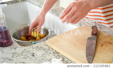 In the contemporary ambiance of a modern kitchen, a young man engages in dinner preparations. His current activity entails meticulously slicing small rainbow potatoes in half on a wooden cutting board In the contemporary ambiance of a modern kitchen, a young man engages in dinner preparations. His current activity entails meticulously slicing small rainbow potatoes in half on a wooden cutting board 112185861