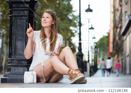 Girl sitting near lamp post using phone to find address Girl sitting near lamp post using phone to find address 112186186