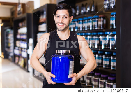 Active muscular man showing his power and holding pot of sport nutrition in shop 112186547