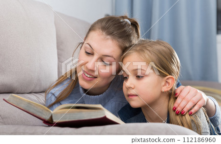 Happy mother with her daughter reading a book on the couch at home 112186960