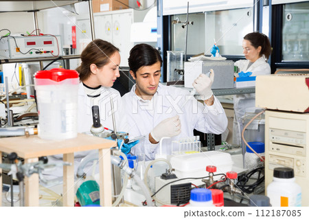 Two young scientists working together in laboratory writing report on results of chemical experiments in notebook 112187085