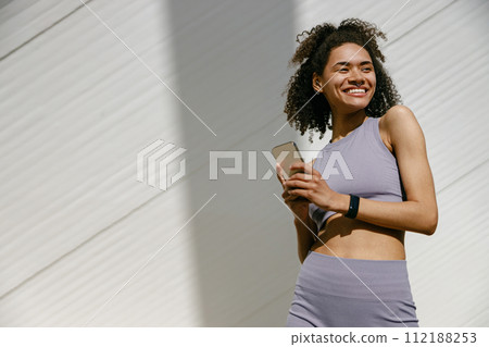 Young smiling woman in sportswear is resting after a workout outside while standing with smartphone Young smiling woman in sportswear is resting after a workout outside while standing with smartphone 112188253