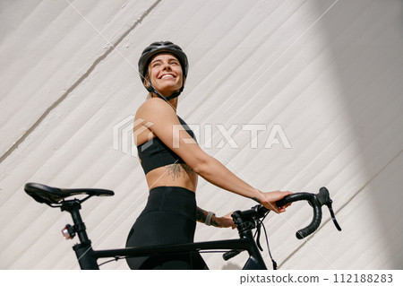 Woman cyclist in protective gear standing with her bike while training outdoors and looks away Woman cyclist in protective gear standing with her bike while training outdoors and looks away 112188283