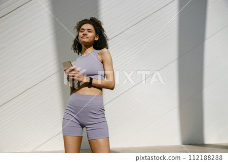Young smiling woman in sportswear is resting after a workout outside while standing with smartphone Young smiling woman in sportswear is resting after a workout outside while standing with smartphone 112188288