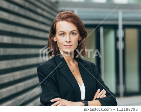 Confident middle-aged woman with crossed arms looking at the camera. Female in black formal clothes posing outdoors. Confident middle-aged woman with crossed arms looking at the camera. Female in black formal clothes posing outdoors. 112188798
