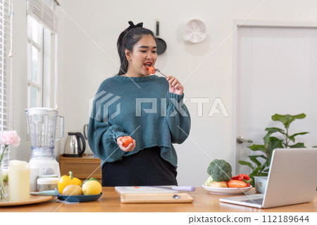 Woman eats a tomato by the laptop on the table by the window in the wood house Woman eats a tomato by the laptop on the table by the window in the wood house 112189644