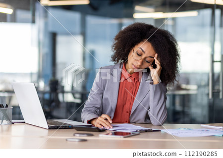 A professional corporate setting capturing a businesswoman at her desk surrounded by documents, a laptop, and a smartphone, seemingly dealing with a stressful workload. 112190285