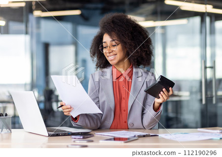 A confident, professional businesswoman is engaged in reviewing documents while working on a laptop at her contemporary office space, demonstrating focus and efficiency. A confident, professional businesswoman is engaged in reviewing documents while working on a laptop at her contemporary office space, demonstrating focus and efficiency. 112190296