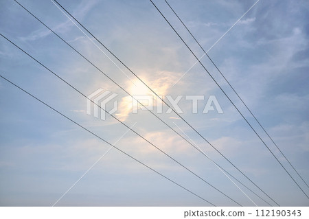 Diagonal hanging high-voltage transmission power lines against blue sky with clouds and shining sun. Empty space. Above-ground electric grid simple background. Diagonal hanging high-voltage transmission power lines against blue sky with clouds and shining sun. Empty space. Above-ground electric grid simple background. 112190343