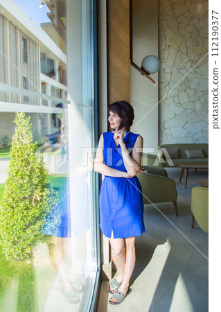 A bright brunette in blue clothes poses by a huge window A bright brunette in blue clothes poses by a huge window 112190377