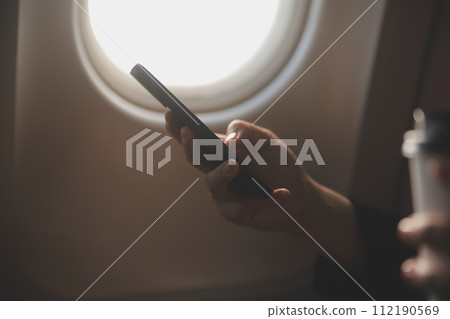 Blonde female tourist checking incoming notification on smartphone sitting on seat of airplane with netbook.Young businesswoman share media from telephone on laptop computer during plane flight Blonde female tourist checking incoming notification on smartphone sitting on seat of airplane with netbook.Young businesswoman share media from telephone on laptop computer during plane flight 112190569