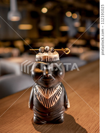 Authentic Tiki Drink Cocktail. Beautiful tropical tiki cocktail on bar counter. Tiki cocktail in monkey mug decorated fig and raspberry on bar in night club or restaurant interior 112191025