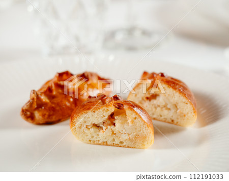 Small russian pies with minced salmon fish Russian Piroshki. Cooked tuna fish hand portioned pies or empanadas cut in half on white plate in elegant restaurant interior. Selective focus, close up. 112191033