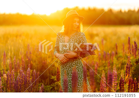 A young woman in a stylish green dress and hat enjoys the sunset in a field with lupins. 112191055