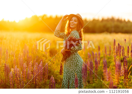 A young woman in a stylish green dress and hat enjoys the sunset in a field with lupins. 112191056