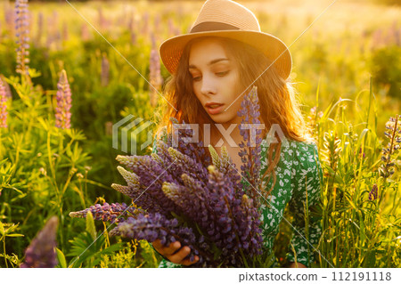 A young woman in a stylish green dress and hat enjoys the sunset in a field with lupins. A young woman in a stylish green dress and hat enjoys the sunset in a field with lupins. 112191118