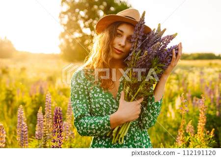 A young woman in a stylish green dress and hat enjoys the sunset in a field with lupins. A young woman in a stylish green dress and hat enjoys the sunset in a field with lupins. 112191120