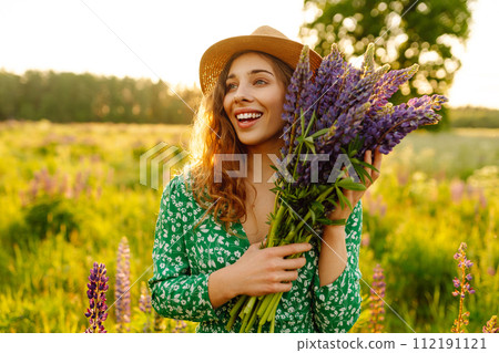 A young woman in a stylish green dress and hat enjoys the sunset in a field with lupins. A young woman in a stylish green dress and hat enjoys the sunset in a field with lupins. 112191121