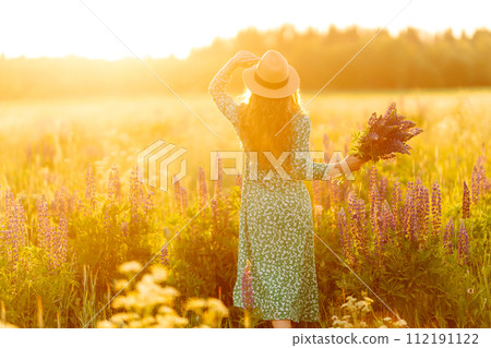 A young woman in a stylish green dress and hat enjoys the sunset in a field with lupins. A young woman in a stylish green dress and hat enjoys the sunset in a field with lupins. 112191122