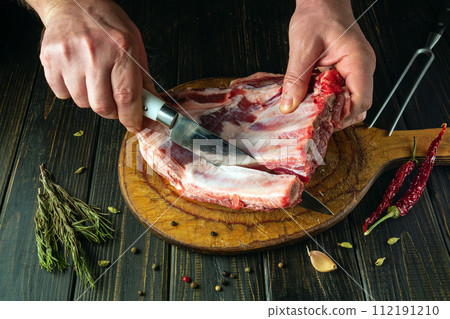 The chef uses a knife to cut the meat from the bone on a kitchen board before preparing the dish. The process of cooking meat on the kitchen table by the hands of a butcher The chef uses a knife to cut the meat from the bone on a kitchen board before preparing the dish. The process of cooking meat on the kitchen table by the hands of a butcher 112191210